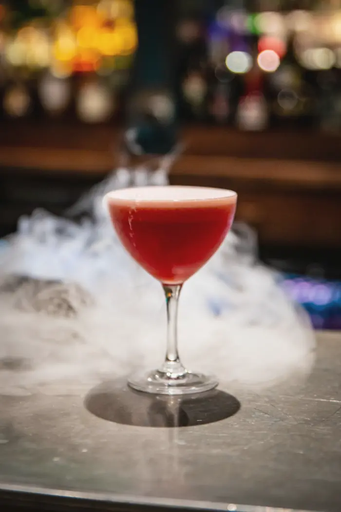 A deep red cocktail in a coupe on a cloud of dry-ice mist at the bar